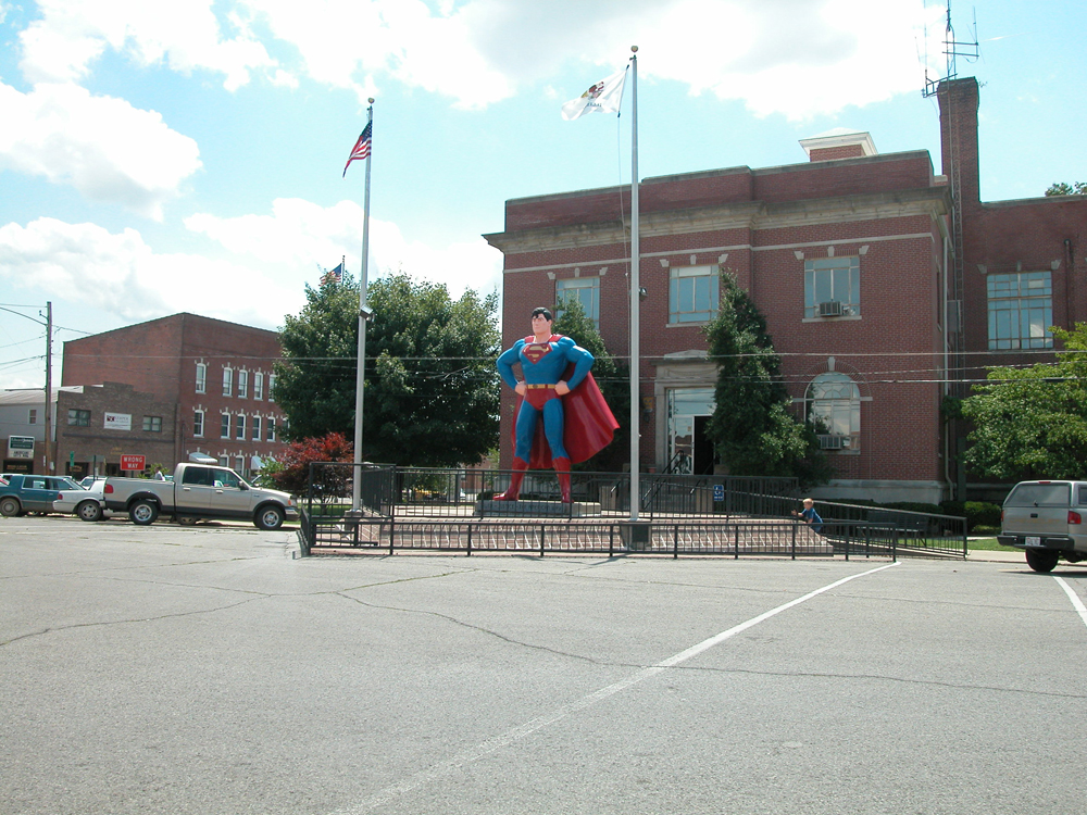 A statue of Superman between two flagpoles and in front of a two-story brick building is shown.