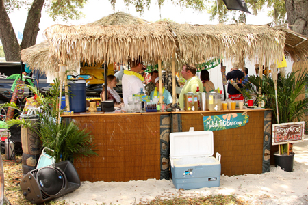 Several people in colorful T-shirts and leis are shown talking and drinking in an outdoor tiki bar setting.