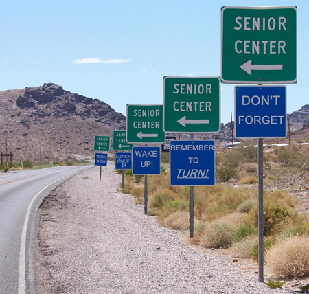 Five sets of road signs, the top one green and the bottom one red in each set, are shown along the right-hand side of a road in a desert setting. The green signs all read “Senior Center” and feature an arrow pointing left. The blue signs, from front to back, read “Don’t Forget,” “Remember to [u]Turn![/u]”, “Wake Up!”, “Lunch Only $4,” and “Turn Now.”