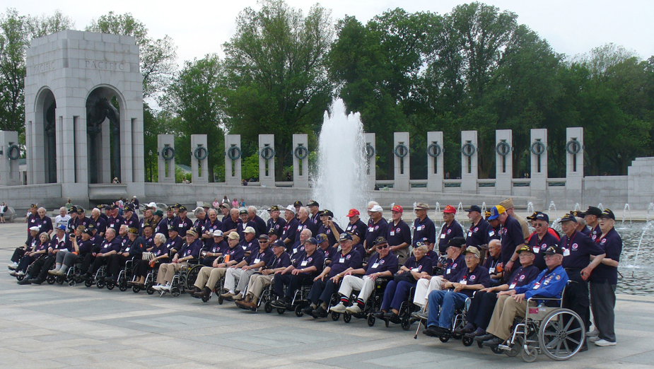 A group of elderly men, many in wheelchairs, all dressed in blue shirts and baseball caps, are shown standing and sitting in a memorial setting, with a fountain and pillars behind them.