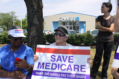 Two elderly women, one holding a red white and blue sign reading “Save Medicare: Make Big Banks Pay Their Share,” are shown sitting under some trees and in front of a suburban bank building. A younger woman, dressed all in black, is shown behind and to the left of the other women.