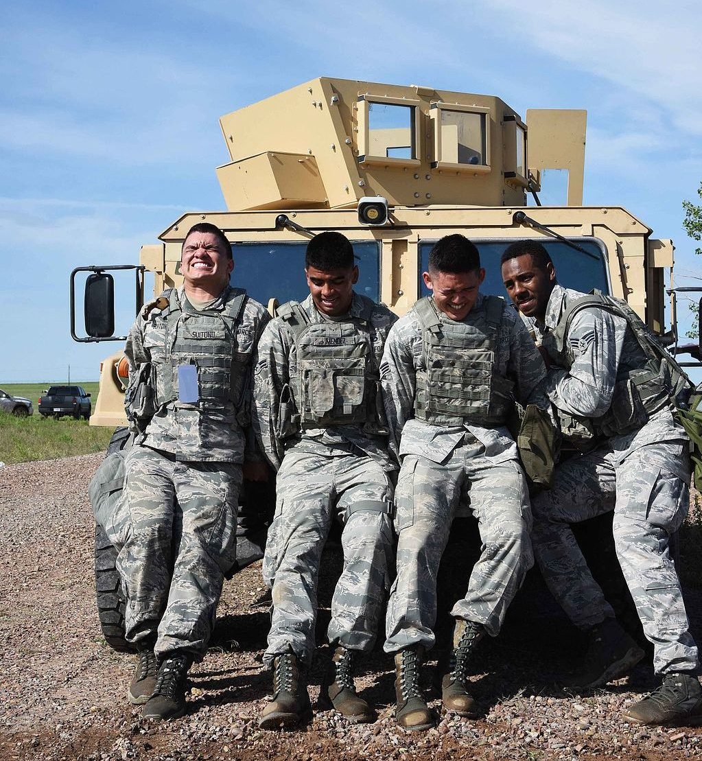 Four soldiers pushing a Humvee. Their backs are against the vehicle and their faces show that they are pushing as hard as they can.