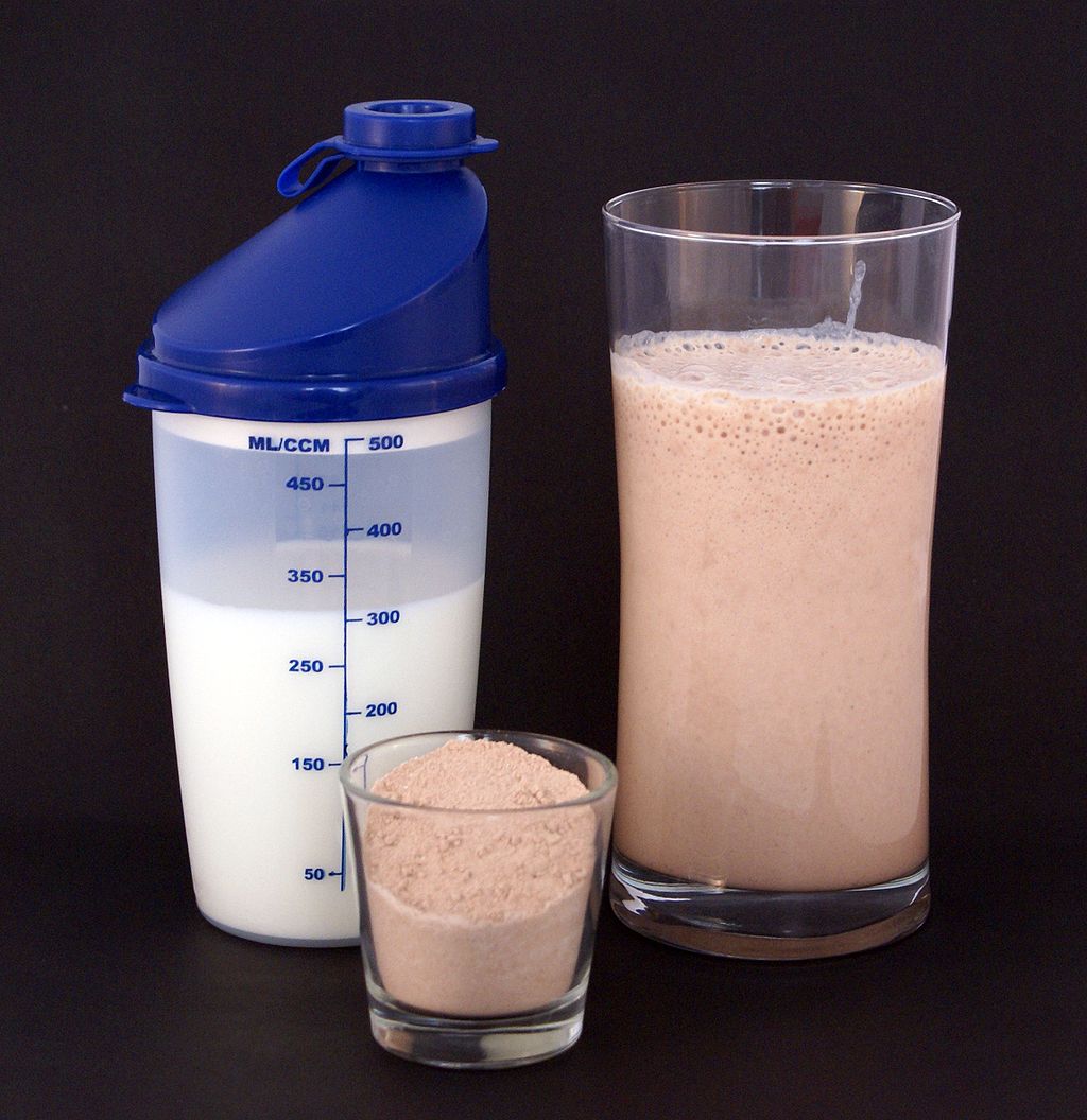 Image shows a glass containing a brown protein shake. Beside the glass are the ingredients used to make the shake: a small container of protein powder and a larger container of milk.