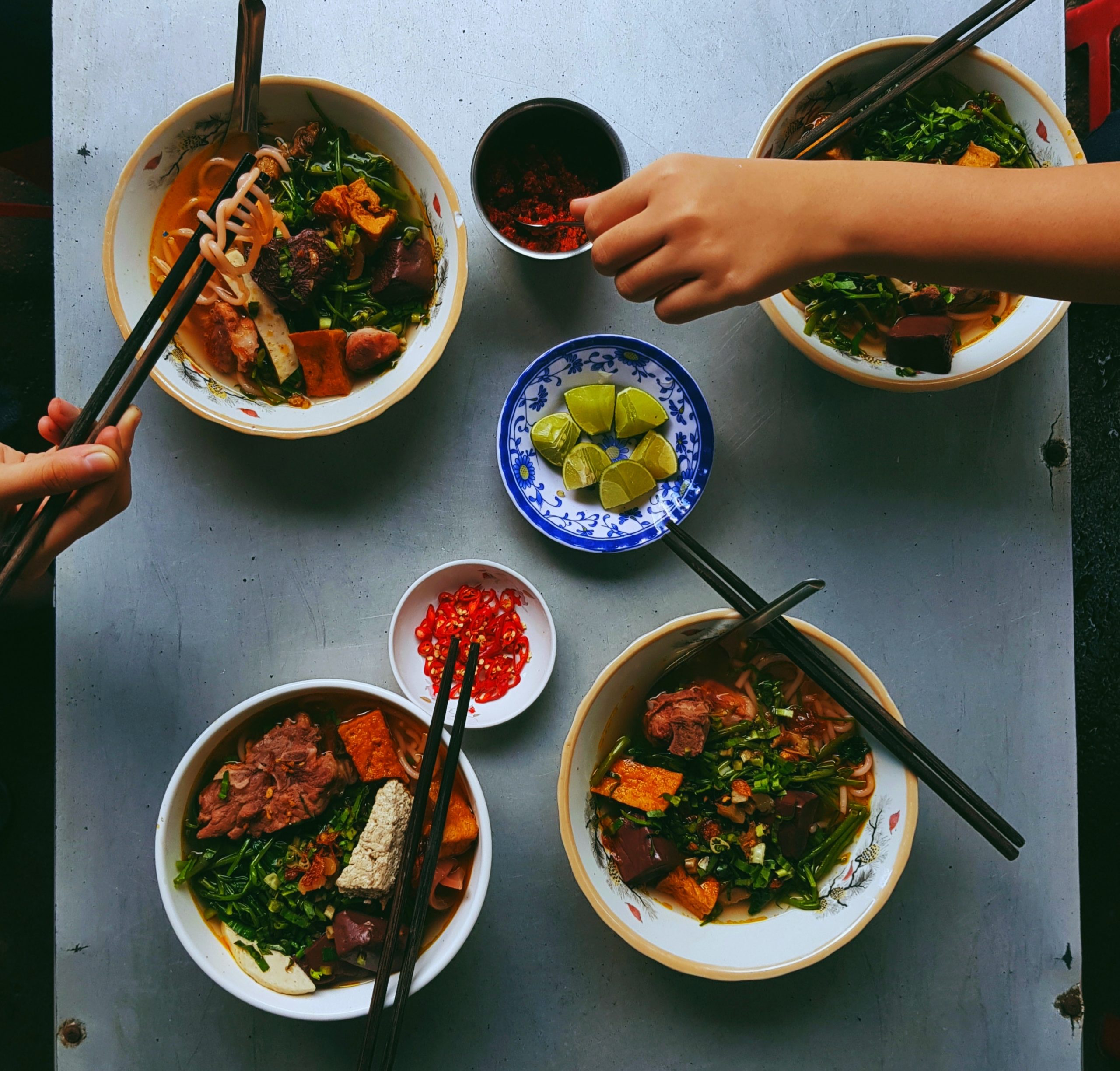 Image shows four bowls of food, each containing noodles, a type of meat, green leafy vegetables and green onions in a broth. Each bowl has chopsticks resting on the side, and there are two smaller bowls in the centre holding lime and chilis.