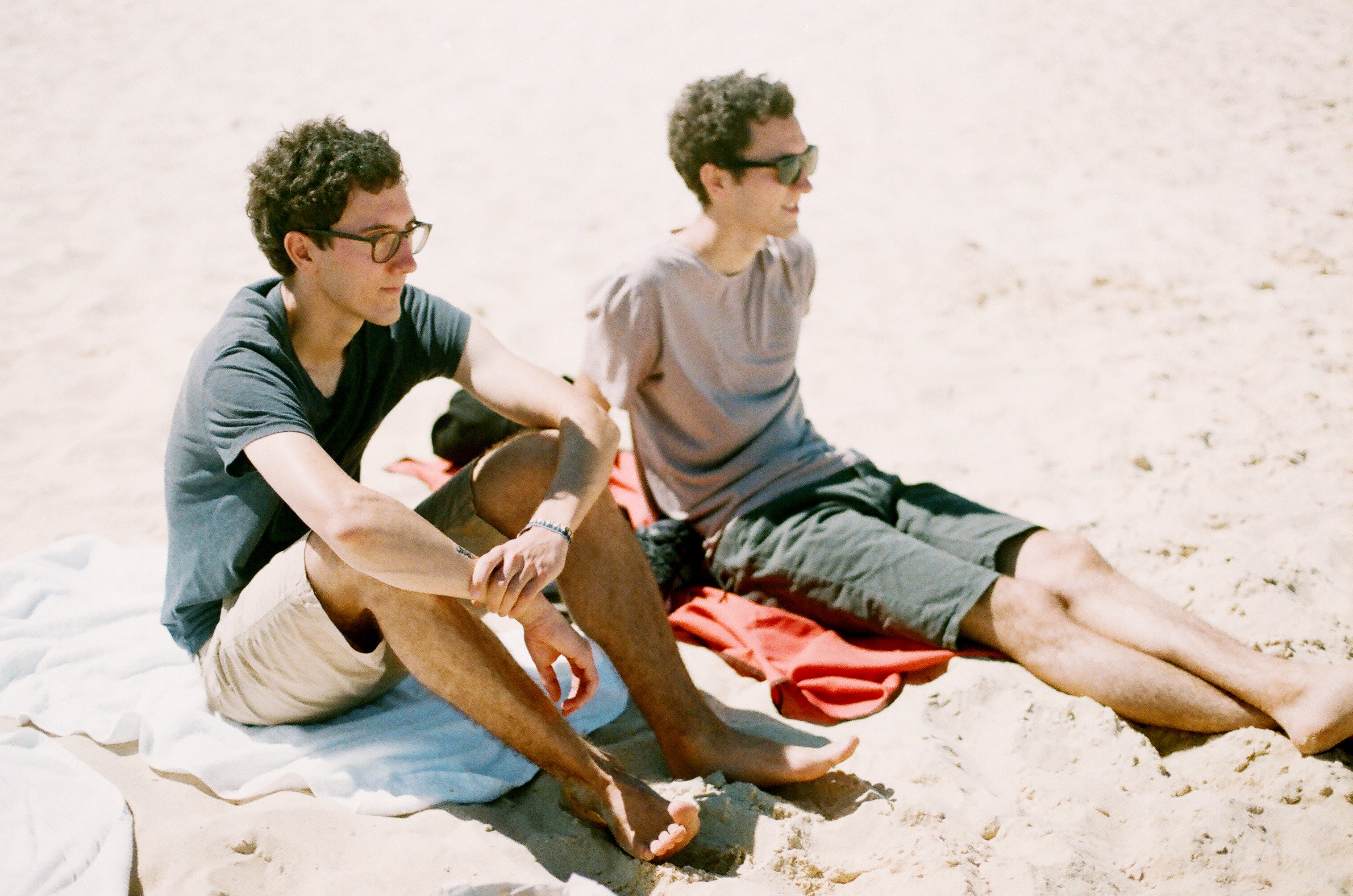 Twins adult males sitting on a beach side by side.