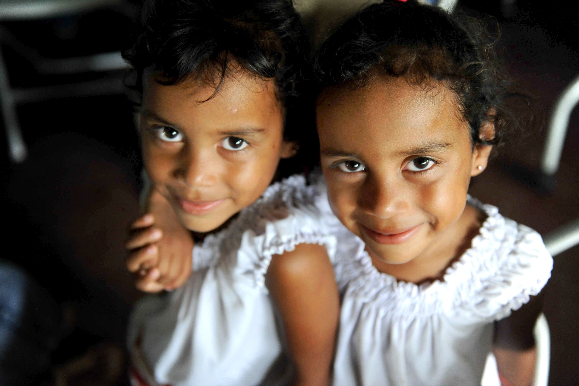 Twin girls with their arms around one another.  Both are wearing white blouses.