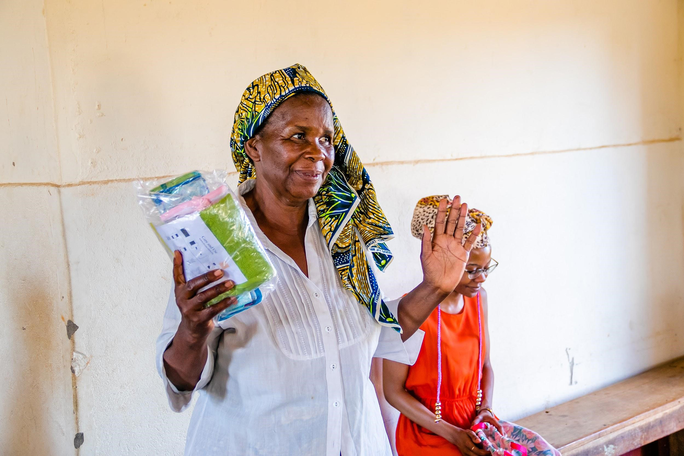 An African woman holding her hands up, holding zip-lock bags and smiling slightly, with another African woman sitting next to her.