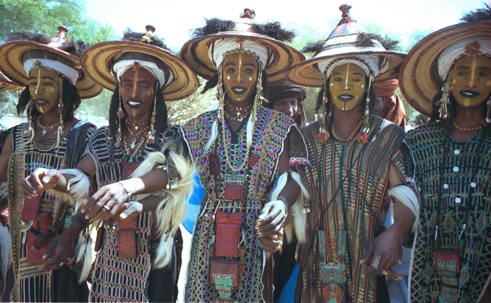 A group of Wodaabe men wearing makeup and elaborate dresses and hats, smiling.