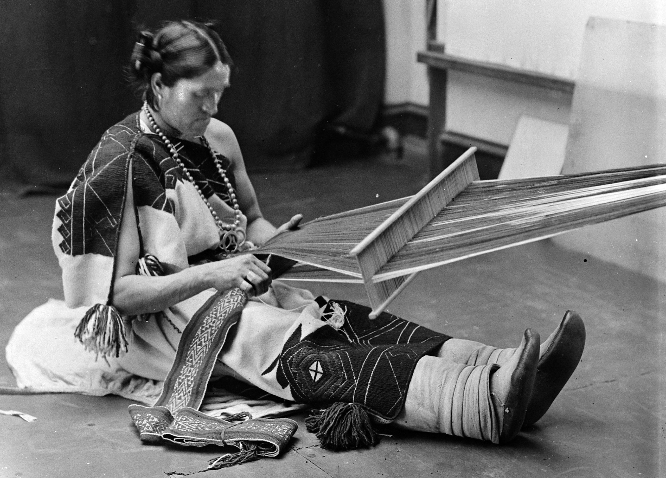 A Zuni Two-Spirit person sitting on the ground weaving cloth, wearing Native clothing.