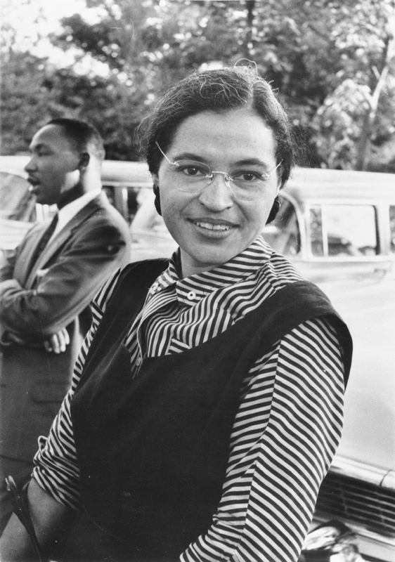 Black and white photo of a young smiling black woman (Rosa Parks) with Martin Luther King talking in the background.