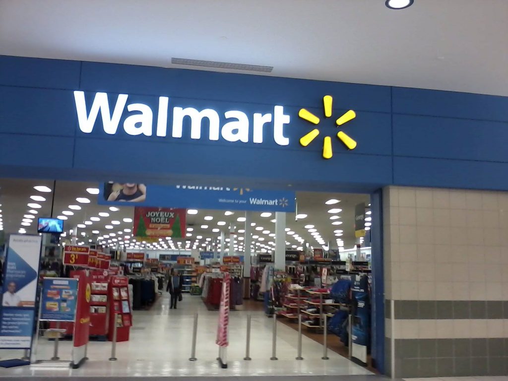 Photo of the entrance to a Walmart store, with a Walmart sign and logo and aisles with products.