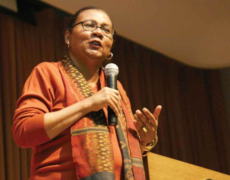 Photo of a black woman in an auditorium, wearing an orange shirt and glasses speaking into a microphone and gesturing.