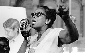 Black and white photo of a black woman speaking passionately into a microphone with her fist raised.