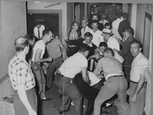 Black and white photo of white men surrounding and attacking a person in a crowded hallway.